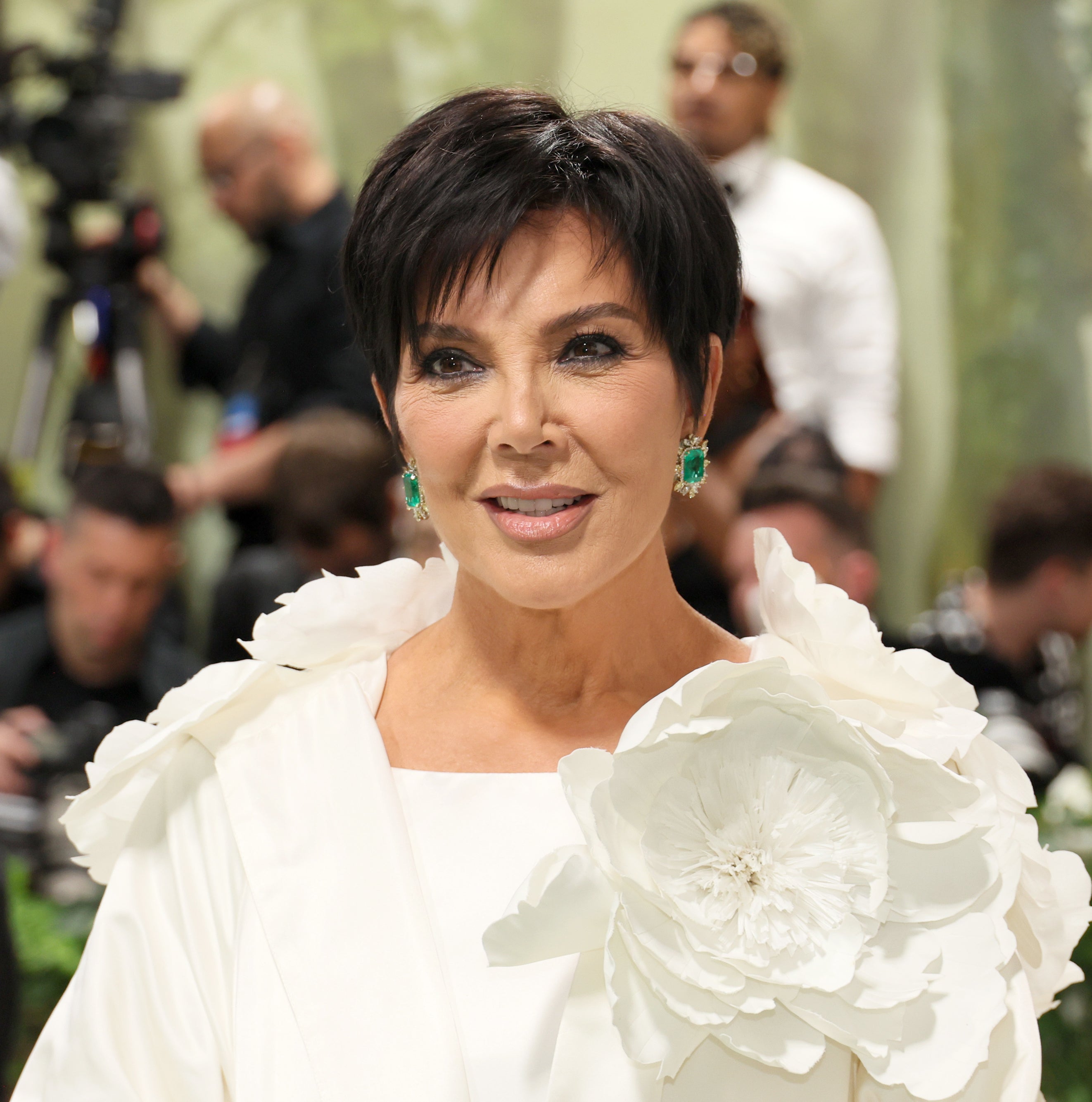 Kris Jenner stands on a red carpet, wearing an outfit adorned with a large floral design on her shoulder