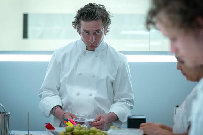 Jeremy Allen White, wearing a chef's uniform, stands in a professional kitchen, focused on work with colleagues nearby