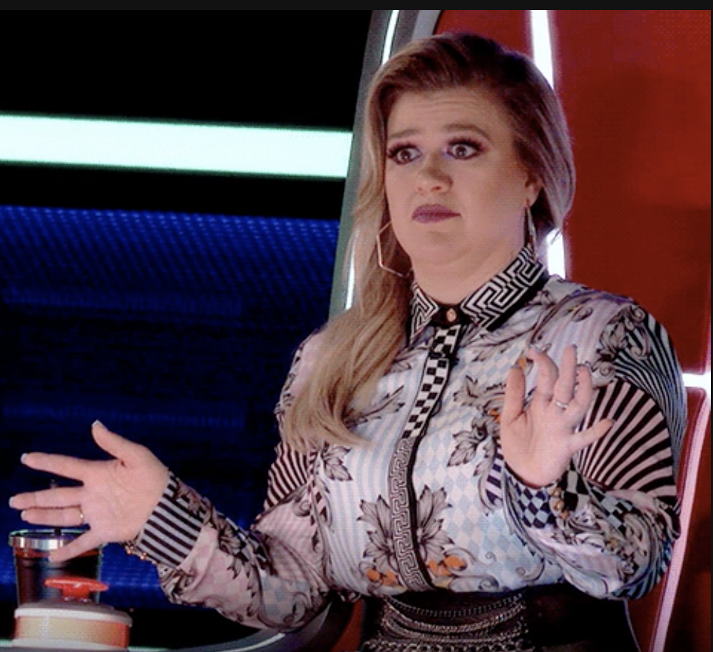 Kelly Clarkson, seated on a set, is gesturing with both hands up in a surprised expression. She is wearing a patterned shirt with stripes and floral designs