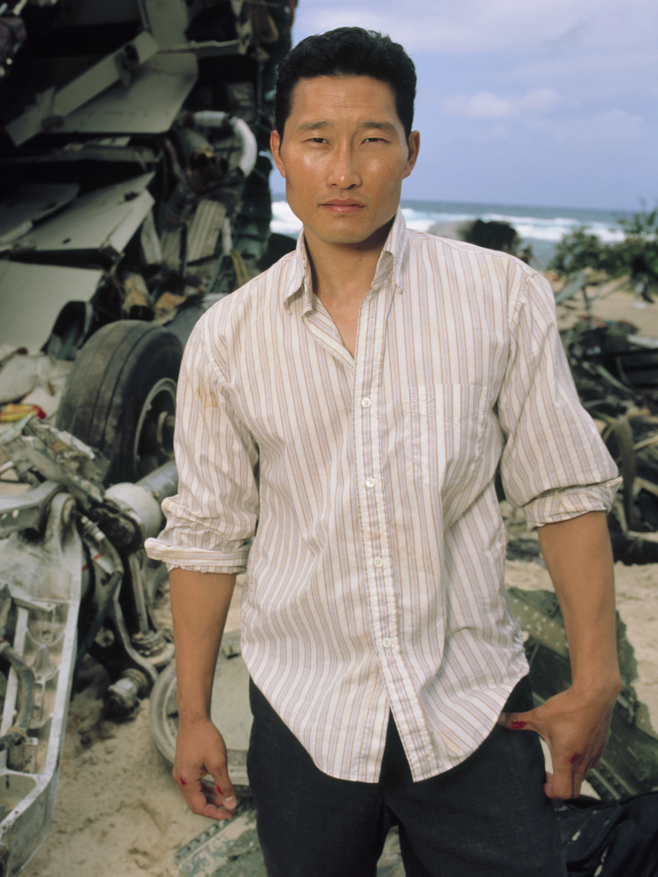 Daniel Dae Kim stands on a beach next to crashed airplane parts, wearing a striped button-down shirt with rolled-up sleeves and dark pants