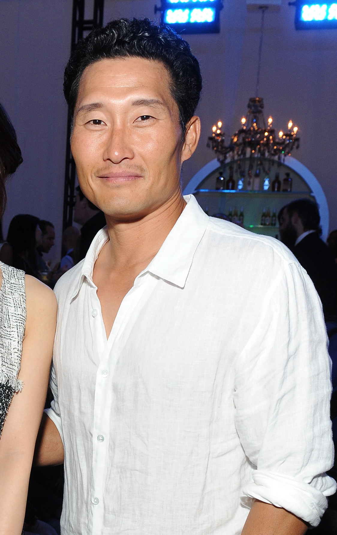 Daniel Dae Kim smiling at an event, wearing a casual white shirt. Background includes people mingling and a chandelier