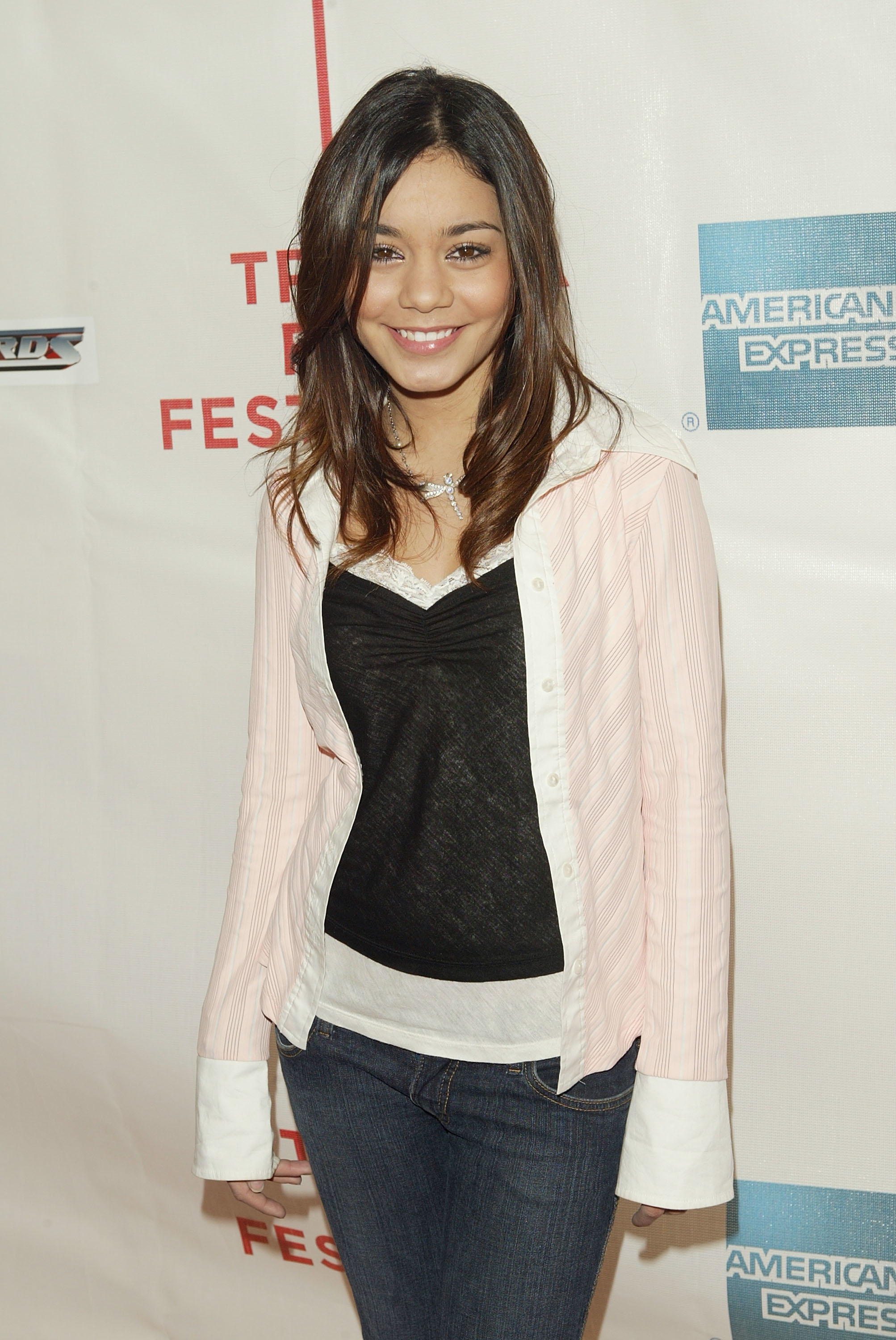 Vanessa Hudgens at an event, wearing a white button-up shirt with pink stripes over a black top and jeans, posing and smiling for the camera