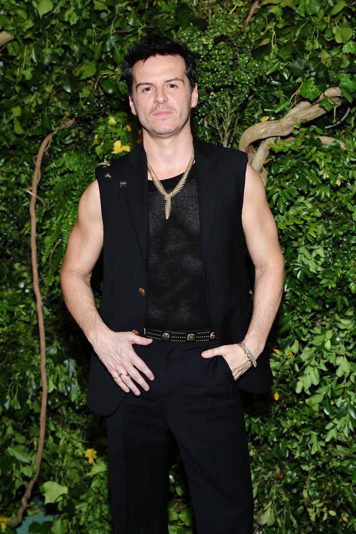 Andrew Scott poses in a sleeveless black vest over a mesh shirt, accessorized with gold necklaces and rings, against a backdrop of lush greenery