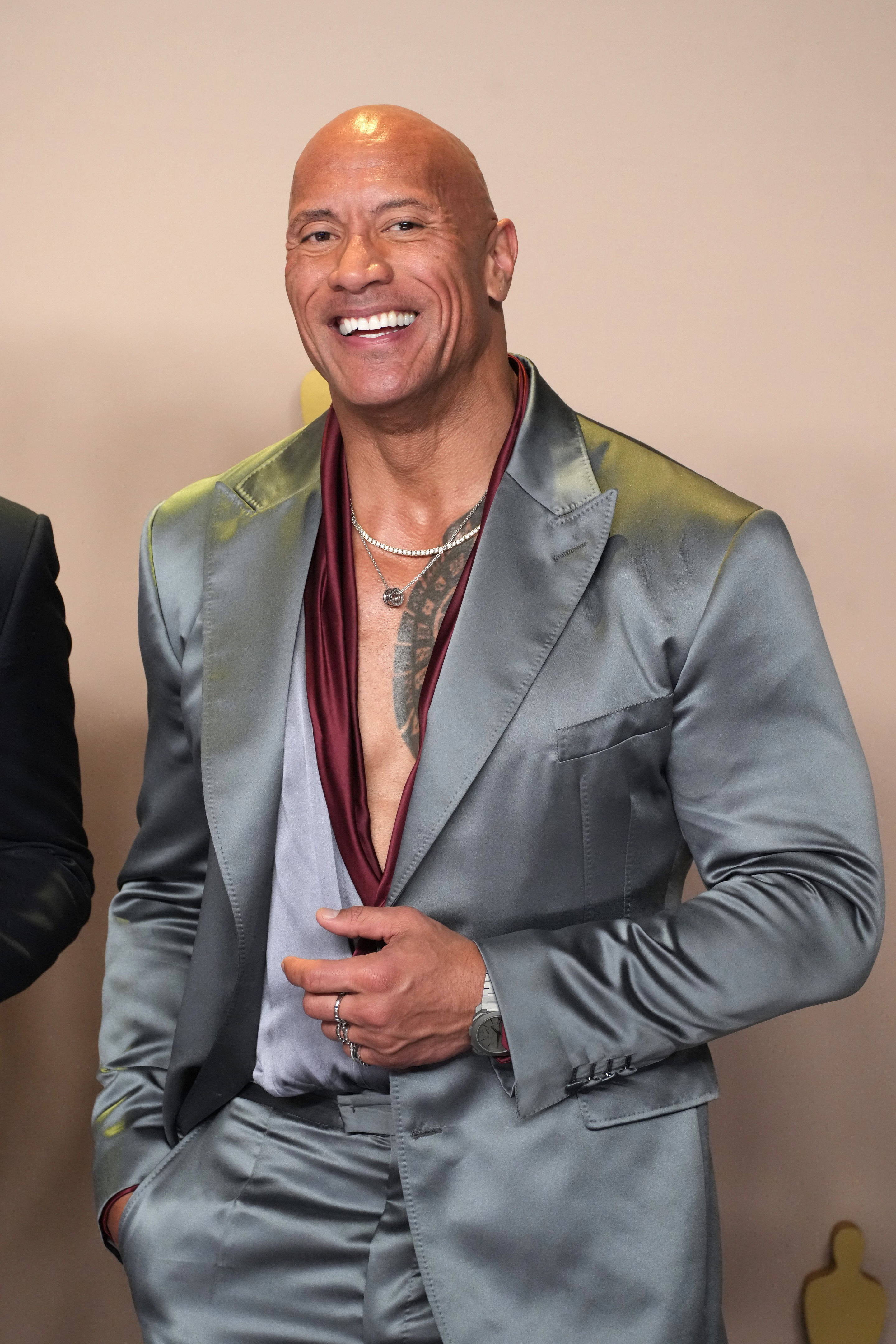 Dwayne Johnson in an open-collar satin suit, smiling, with a tattoo visible on his chest