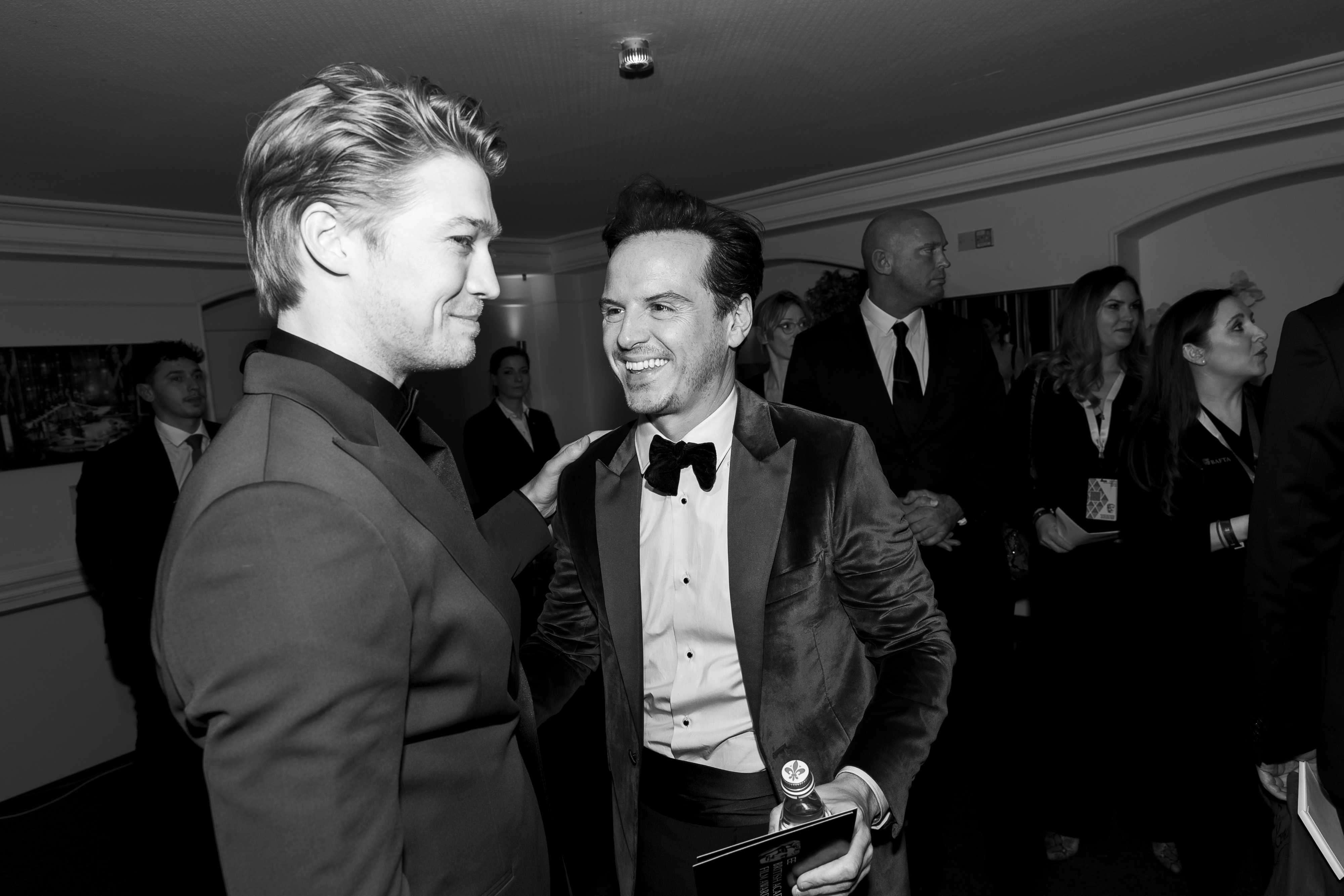 Joe Alwyn and Andrew Scott smiling and talking in a formal setting, wearing suits while surrounded by other guests