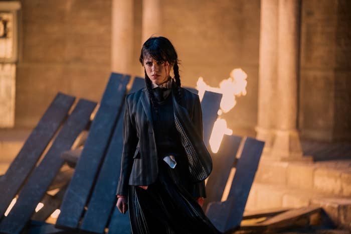 Jenna Ortega as Wednesday Addams in a dark, gothic outfit, standing in front of a fire and collapsed wooden beams in the TV series &quot;Wednesday&quot;