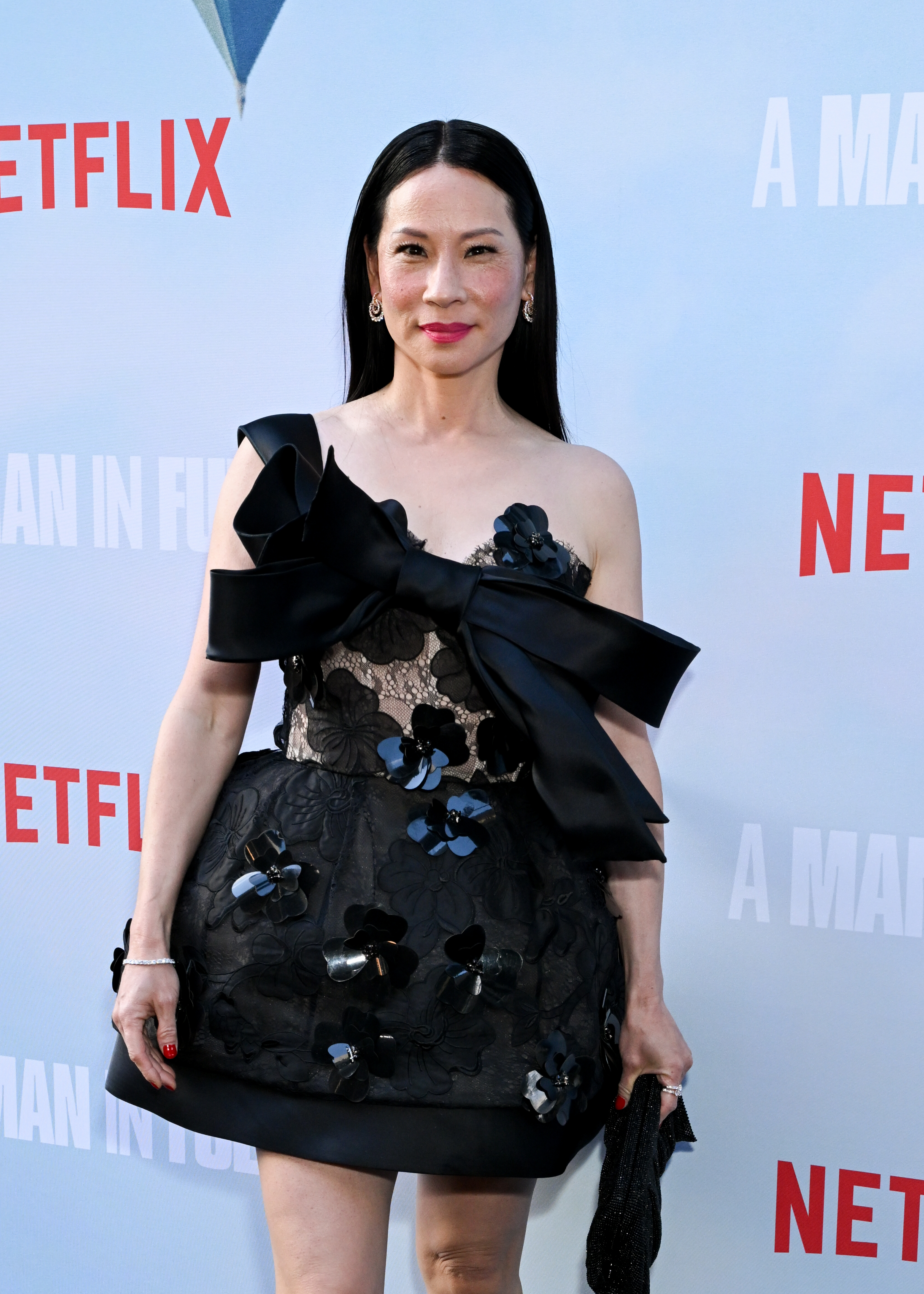 Lucy Liu on the red carpet wearing a black, floral embroidered dress with large bow accents at a Netflix event for "A Man in Full."