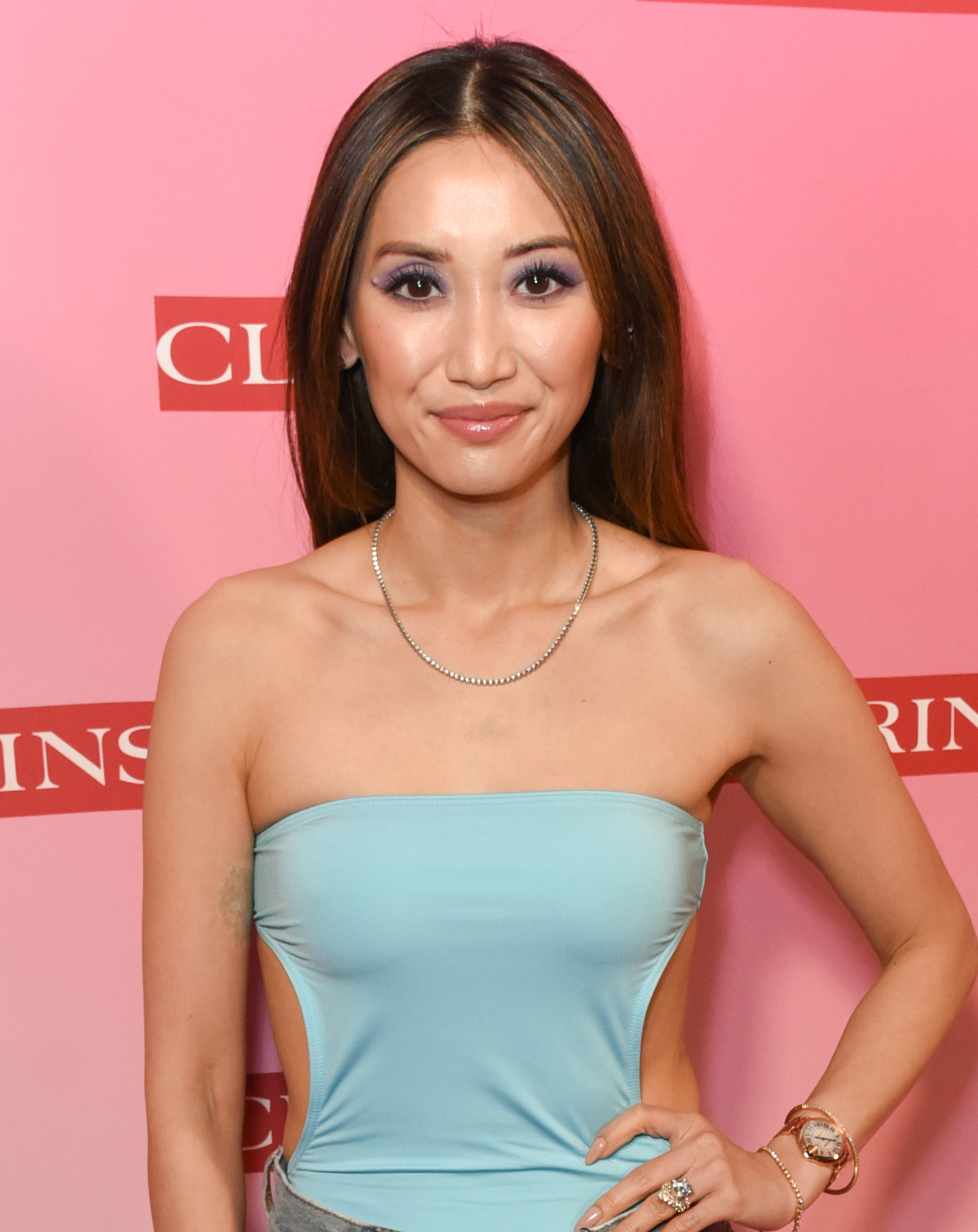 A woman poses for a photo, smiling gently. She is wearing a strapless, light blue dress with cut-outs at the sides, accessorized with simple jewelry