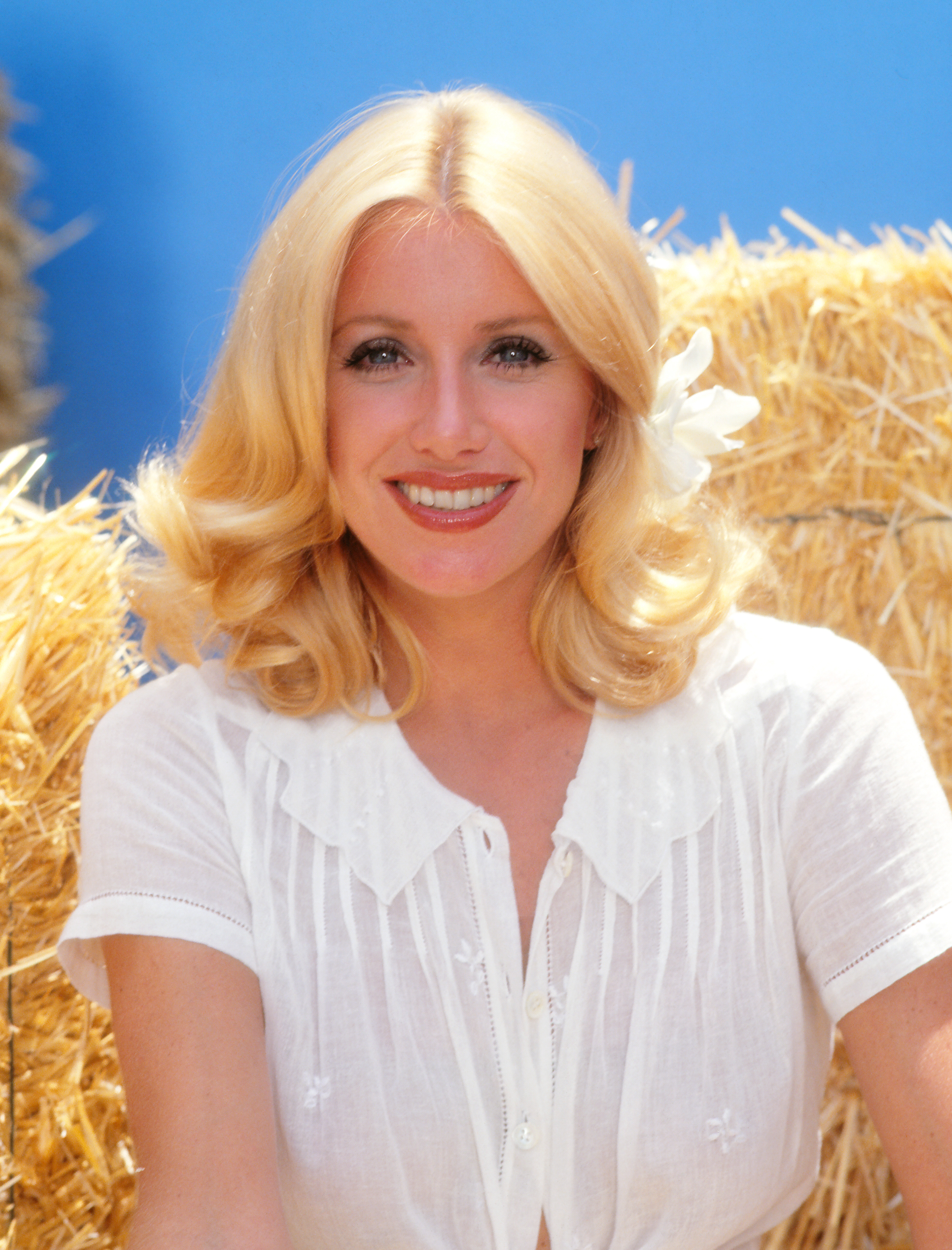 A woman with blonde hair and a white flower in her hair smiles, wearing a white blouse, in front of hay bales