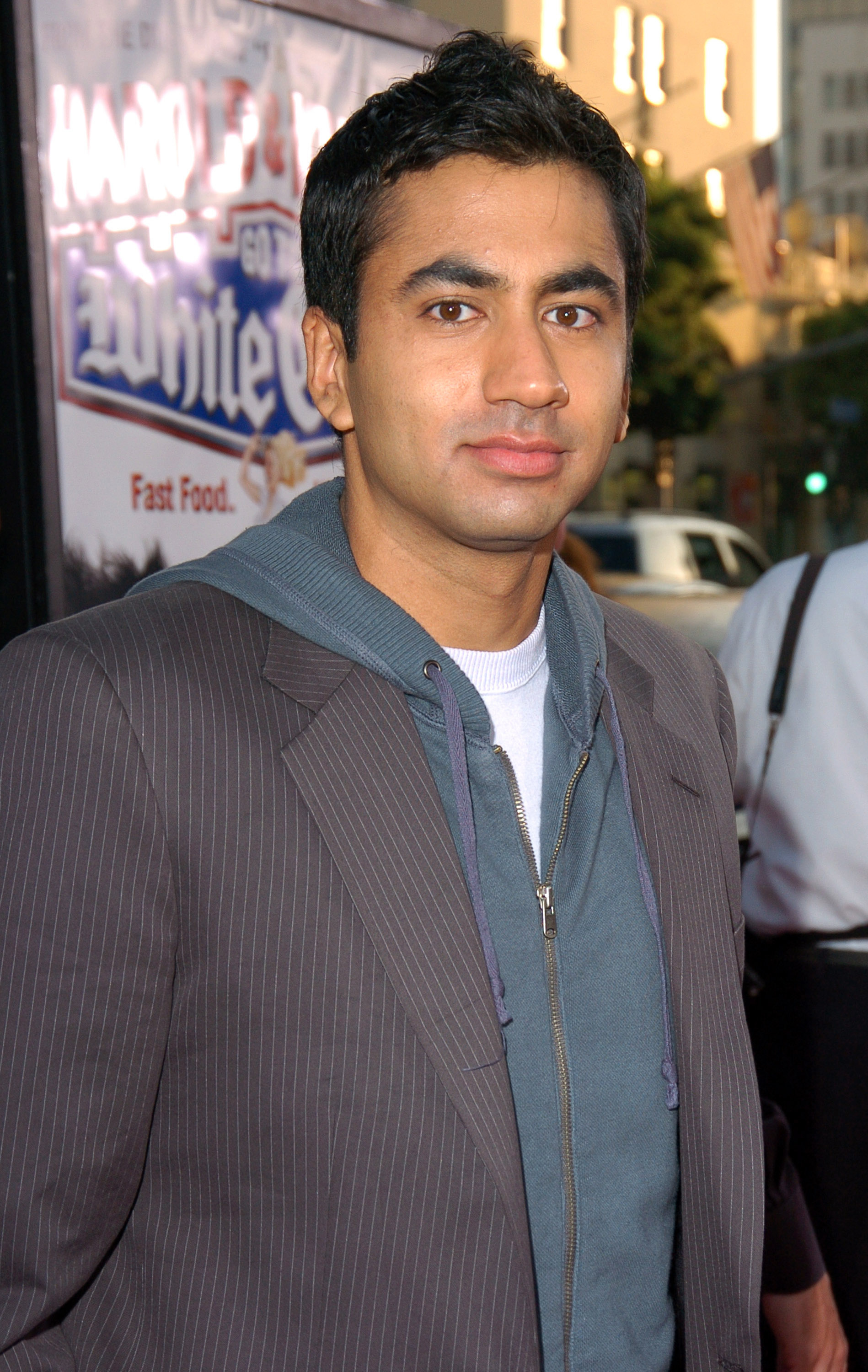 Kal Penn at an event, wearing a striped blazer over a hoodie and t-shirt, standing in front of a promotional poster