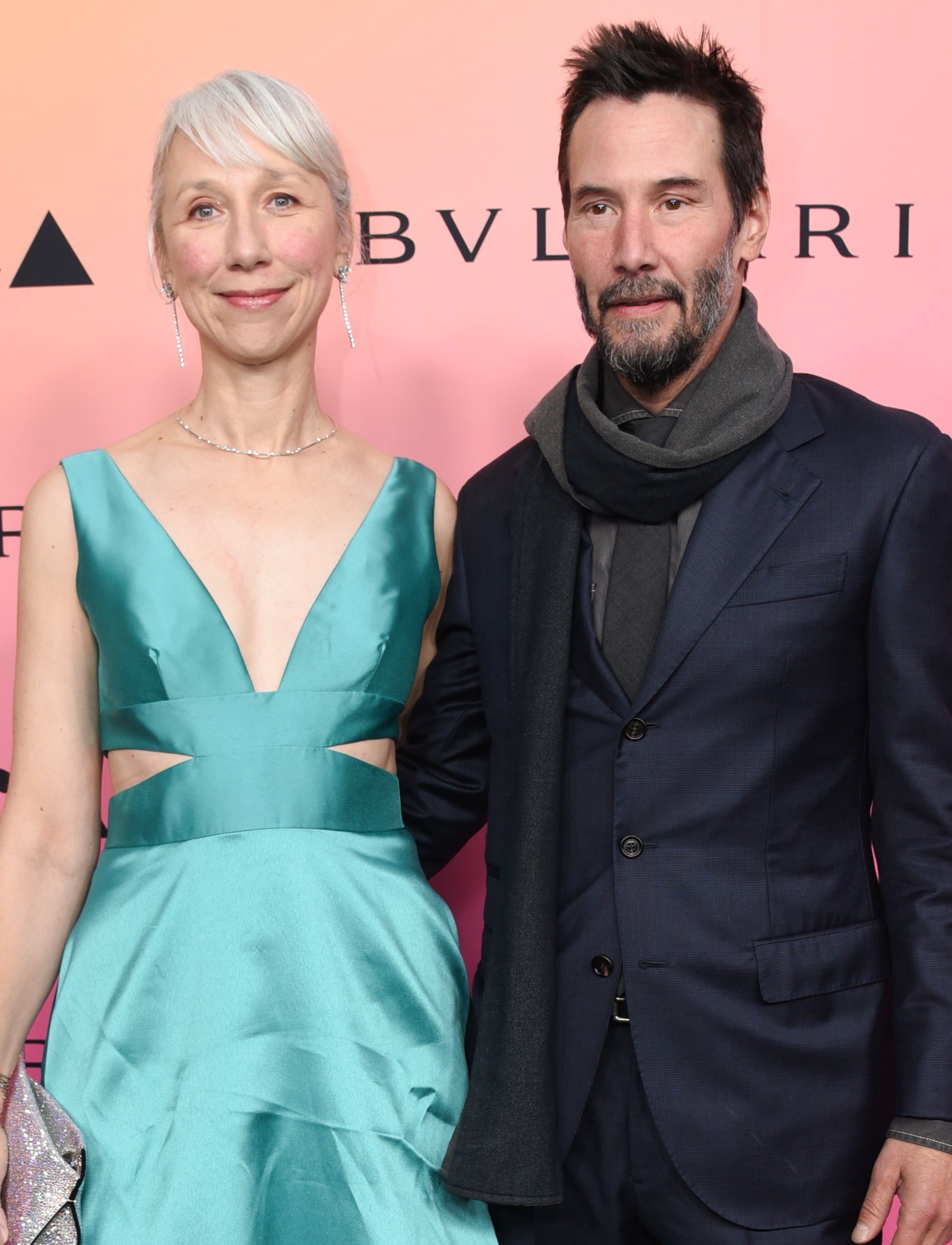 Two people, Alexandra Grant in a satin gown with cutouts, and Keanu Reeves in a dark suit and scarf, pose on the red carpet at an event with BVLGARI signage