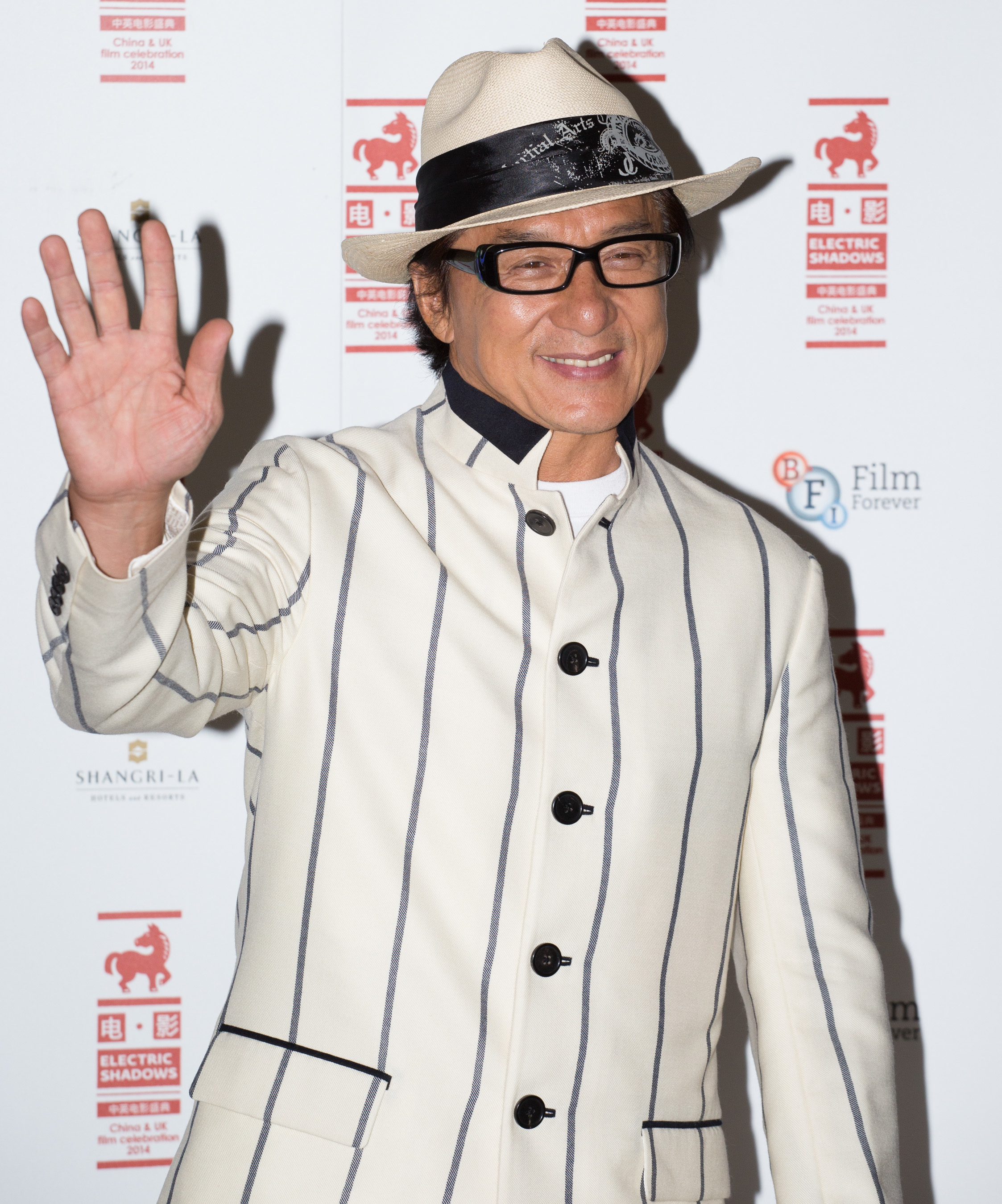 Jackie Chan waves while wearing a white pinstripe suit and a fedora at an event with a step-and-repeat backdrop