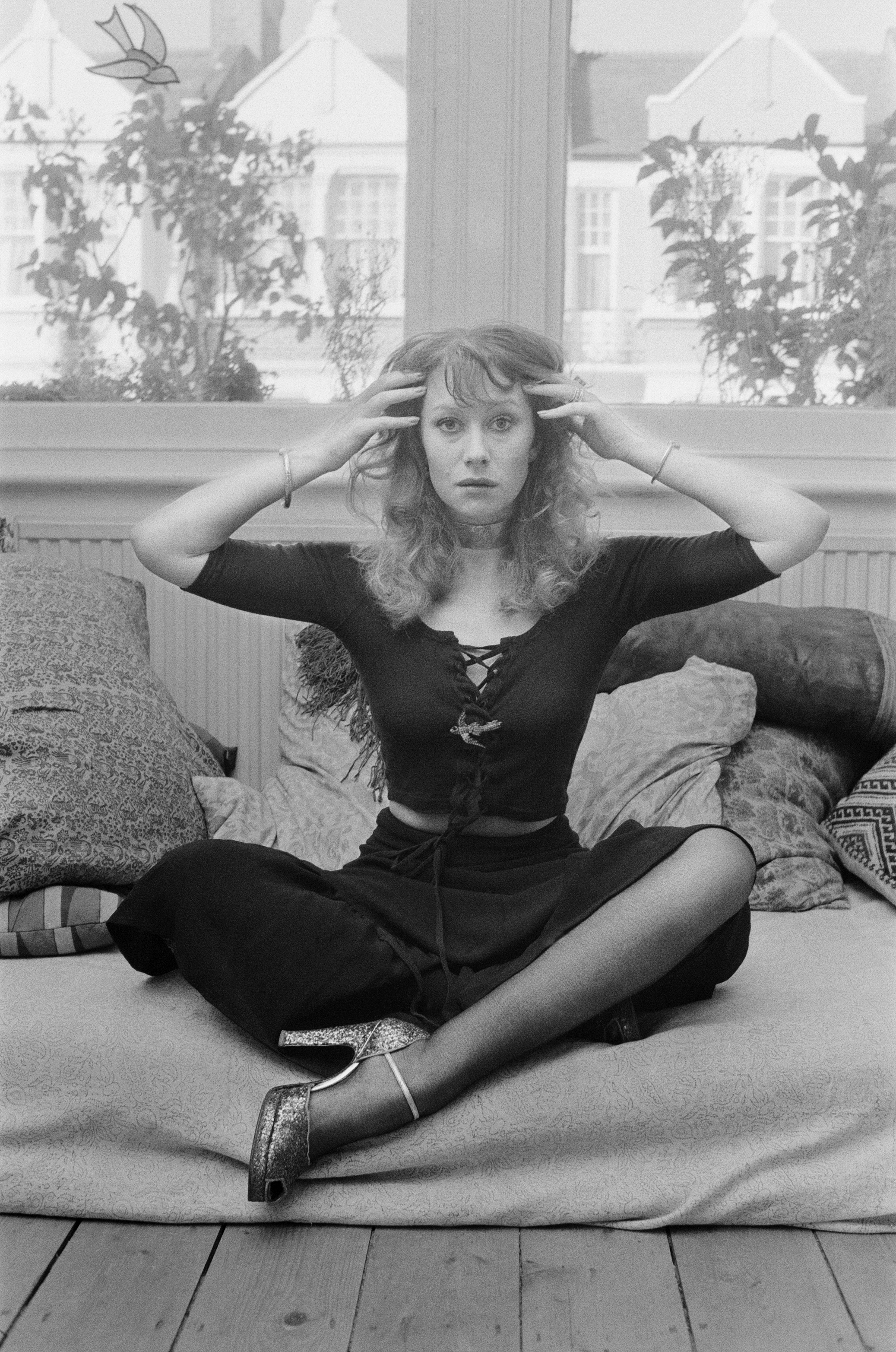 Gwyneth Paltrow sitting cross-legged on a couch, wearing a laced top, skirt, and high-heels, holding her head in a gesture, with a windowed background