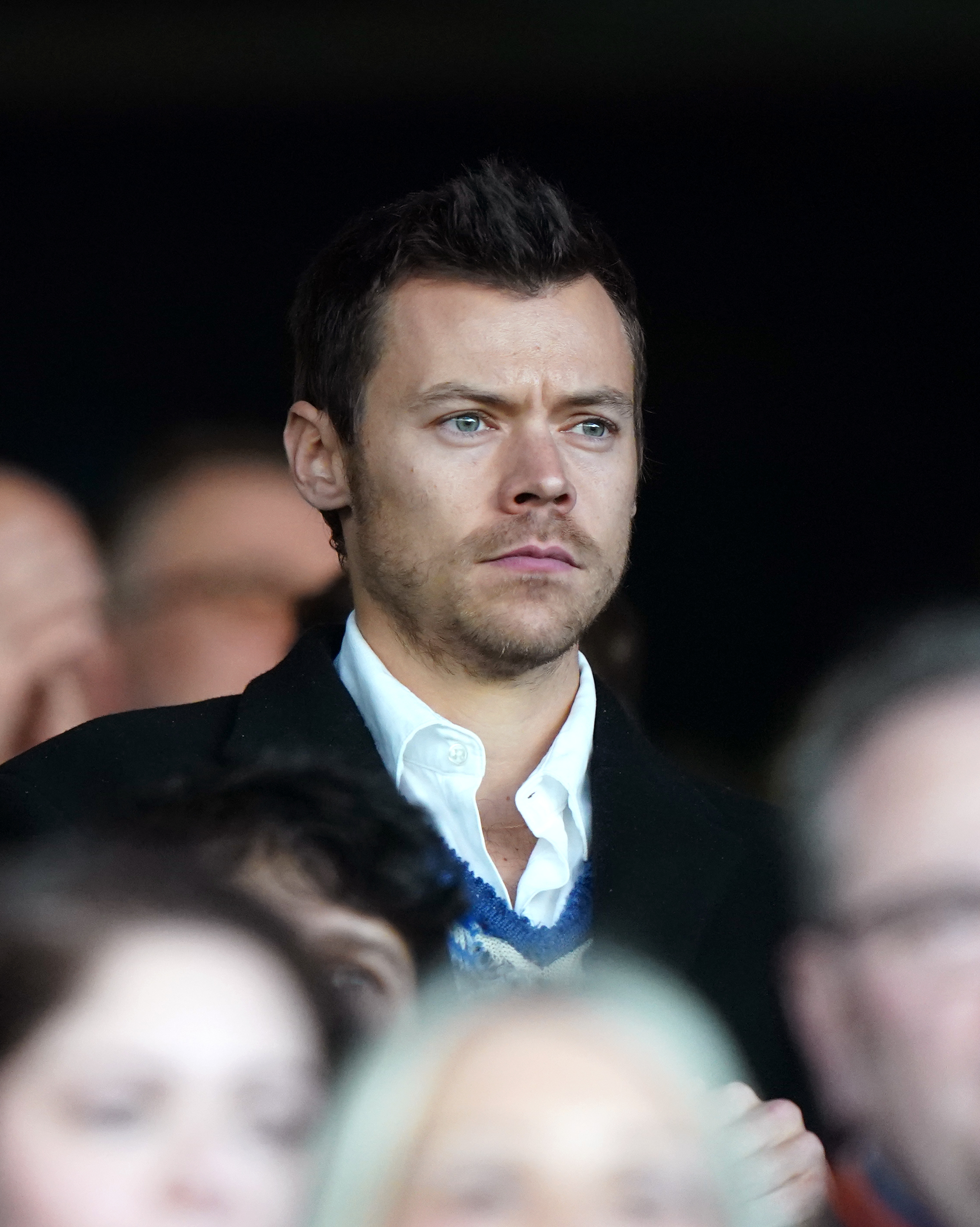 Harry Styles is seen seated at an event, wearing a collared shirt under a sweater and blazer