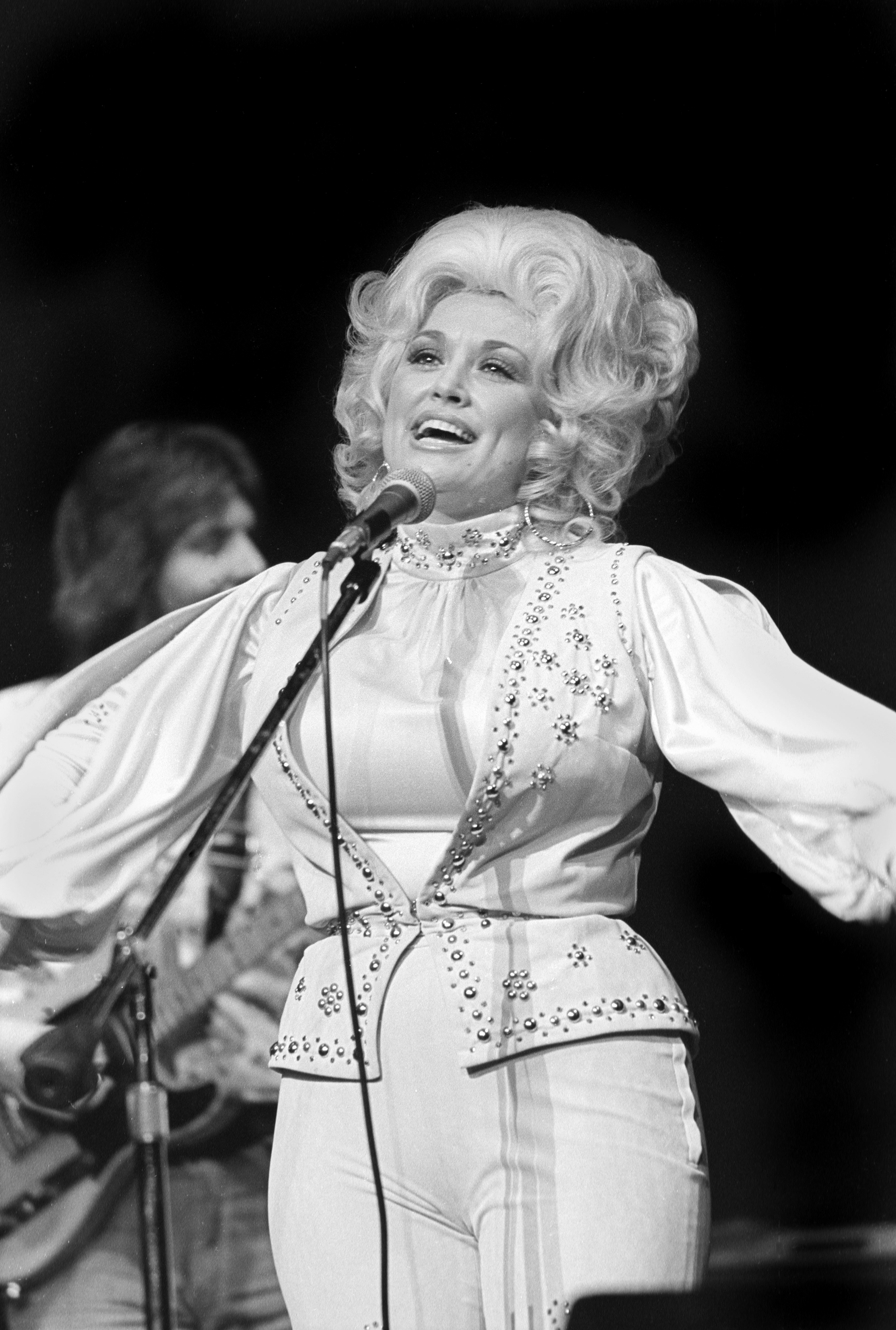 Dolly Parton performing on stage, singing into a microphone while wearing a detailed white outfit with embroidery and puffed sleeves
