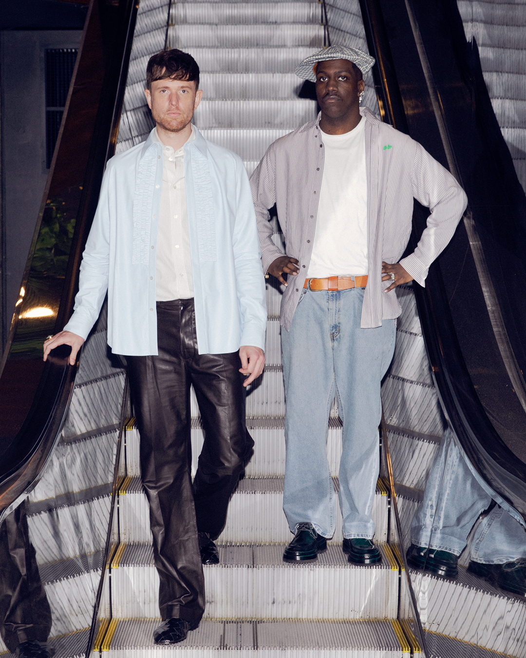 James Blake in a light shirt and leather pants stands next to Lil Yachty in casual attire on an escalator