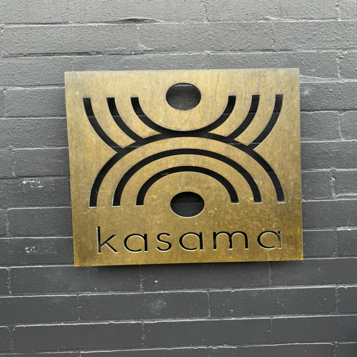 Square sign with geometric design above the word "kasama" mounted on a brick wall