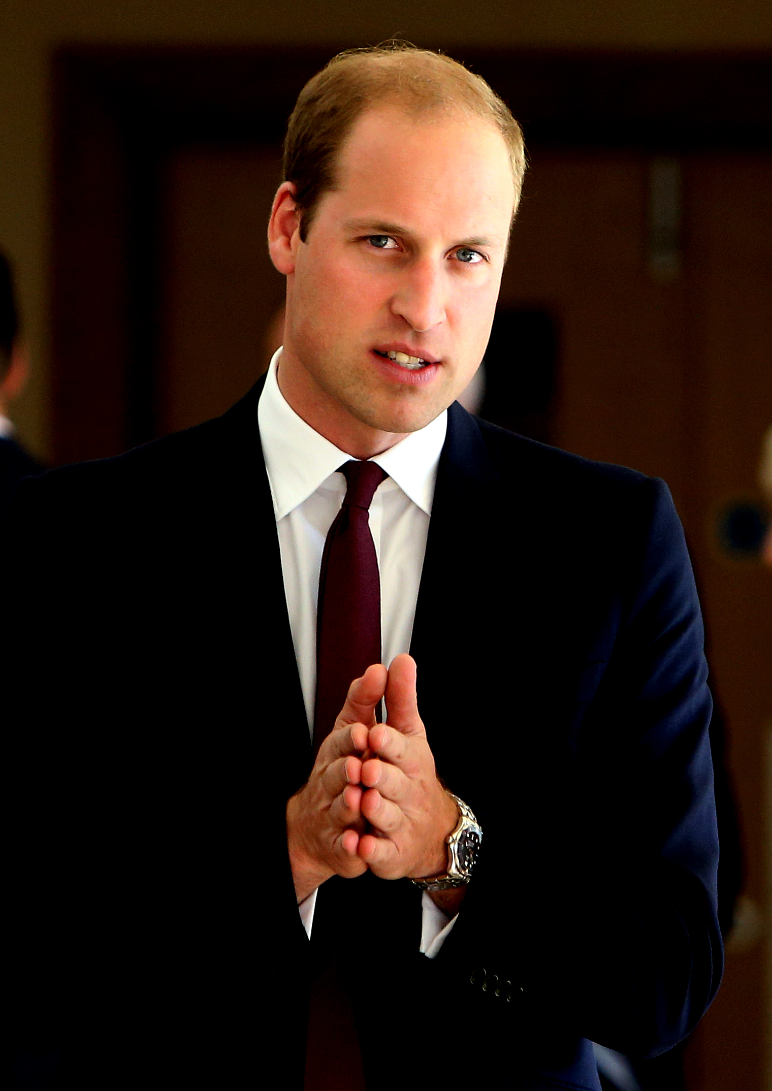 Prince William dressed in a dark suit, white shirt, and maroon tie, standing with hands clasped in a formal setting