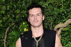 Andrew Scott poses in a sleeveless black vest over a mesh shirt, accessorized with gold necklaces and rings, against a backdrop of lush greenery