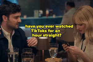 A man and Taylor Swift are sitting at a table in a restaurant. The man is gesturing while Taylor Swift looks at her phone intently