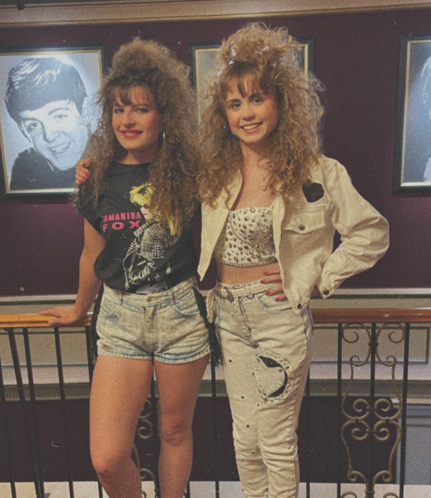 Two unidentified women with 1980s hairstyles and fashion. One wears a graphic T-shirt and shorts, the other wears a leopard print top, white jacket, and distressed pants