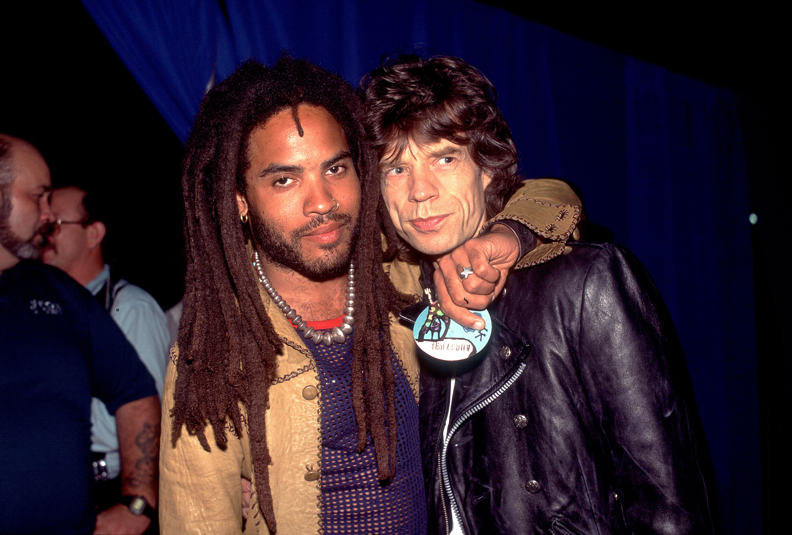 Lenny Kravitz and Mick Jagger pose together; Lenny wears a casual jacket with his arm around Mick, who wears a leather jacket