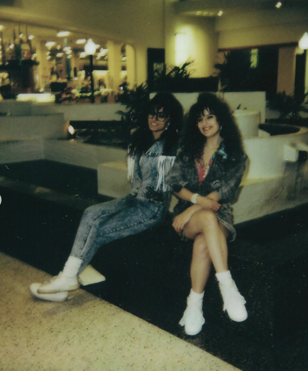 Two women with big hairstyles and 80s fashion, including denim and fringe, sit together in a shopping mall&#x27;s atrium area, smiling at the camera