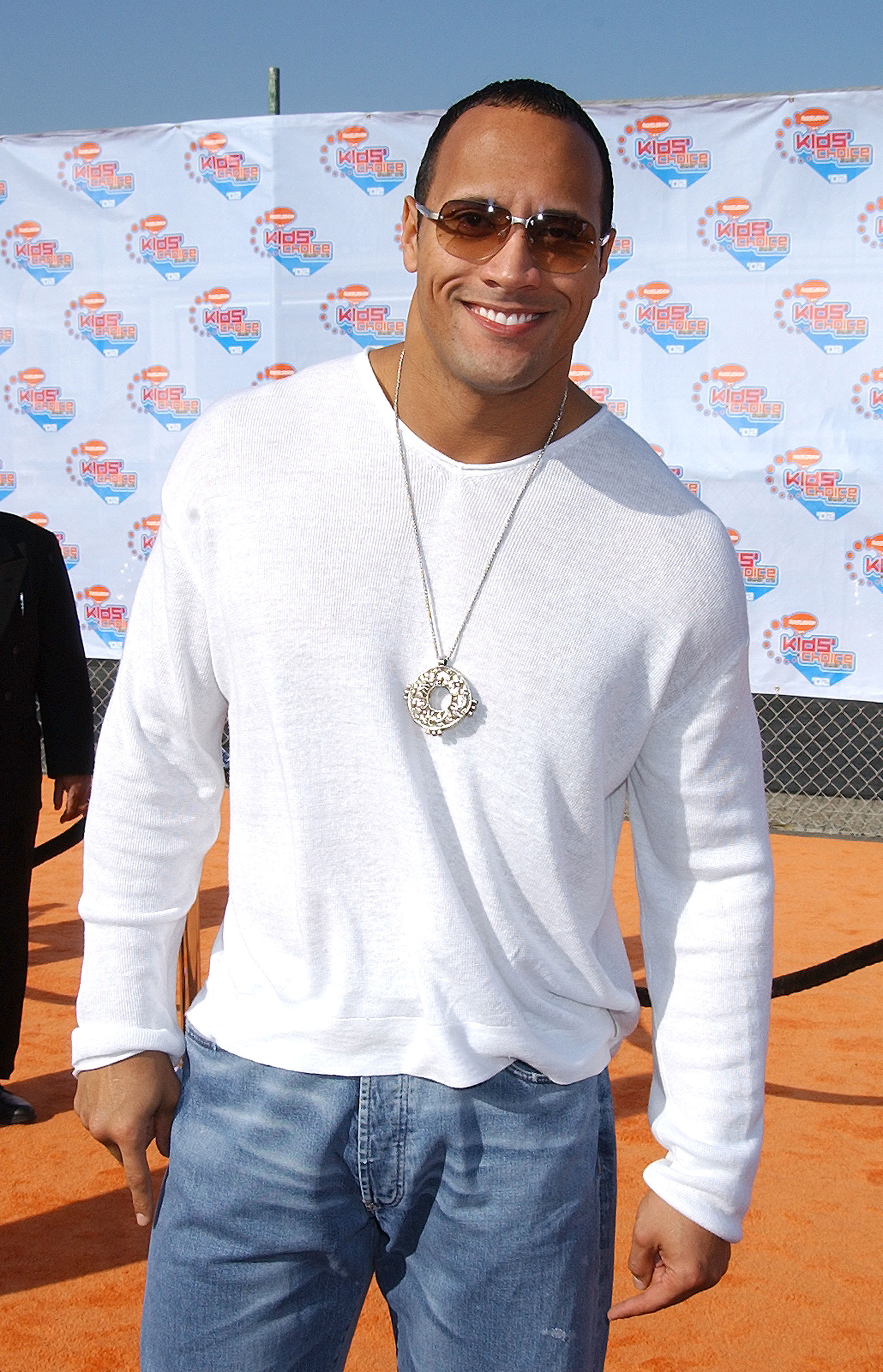 Dwayne Johnson on the red carpet wearing a white long-sleeve shirt, blue jeans, sunglasses, and a silver pendant necklace, smiling at the camera