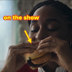 A person with braided hair is happily eating a sandwich, located in an indoor setting