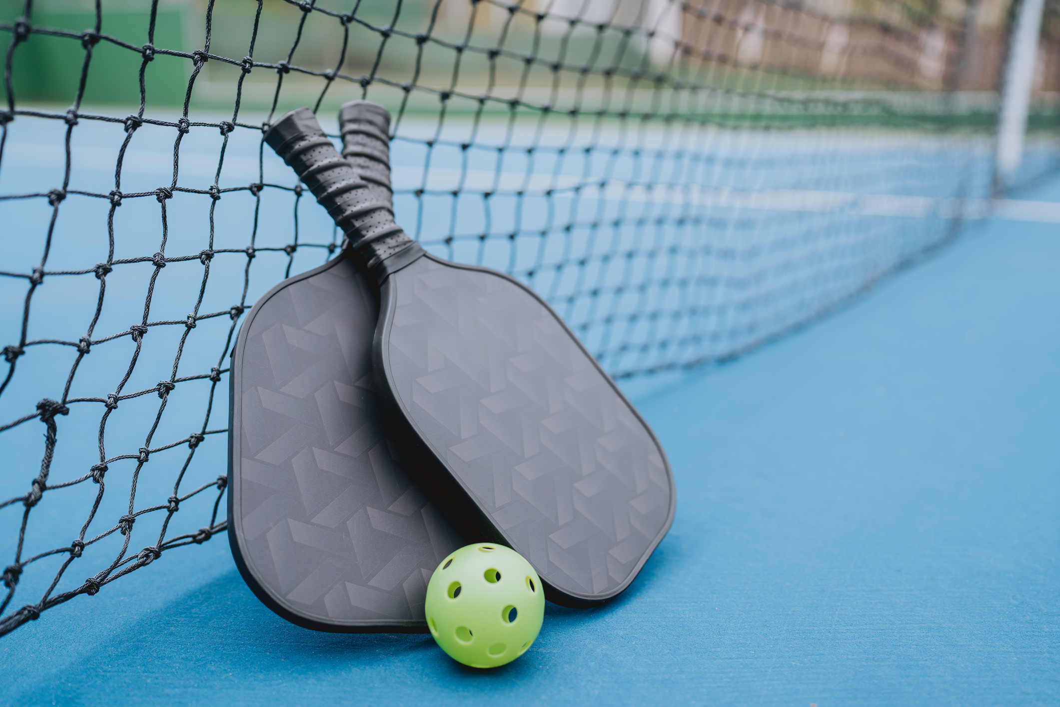 Two pickleball paddles rest against a net, with a bright green plastic ball in front of them on a court