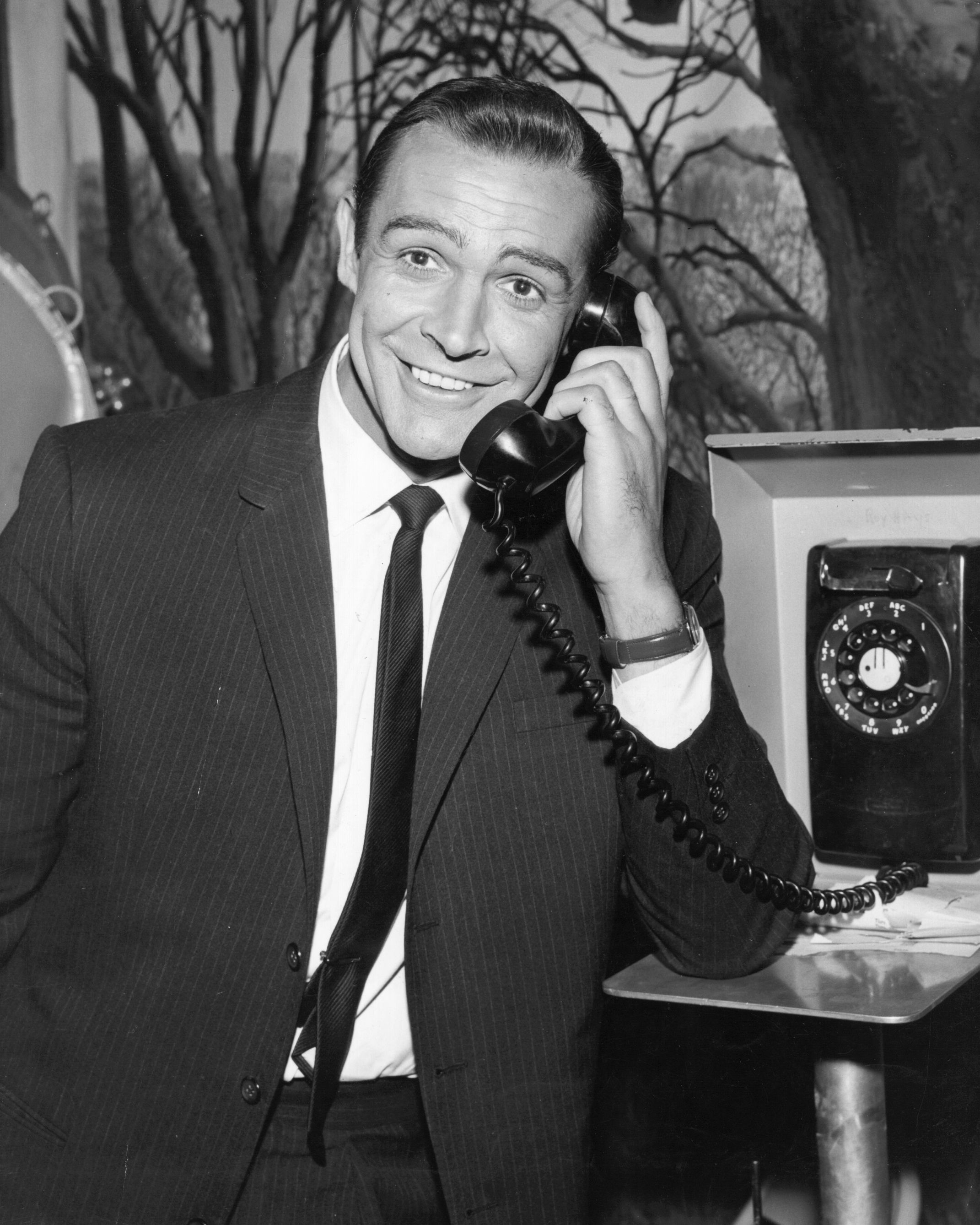 Sean Connery, in a suit and tie, smiling while speaking on a rotary phone