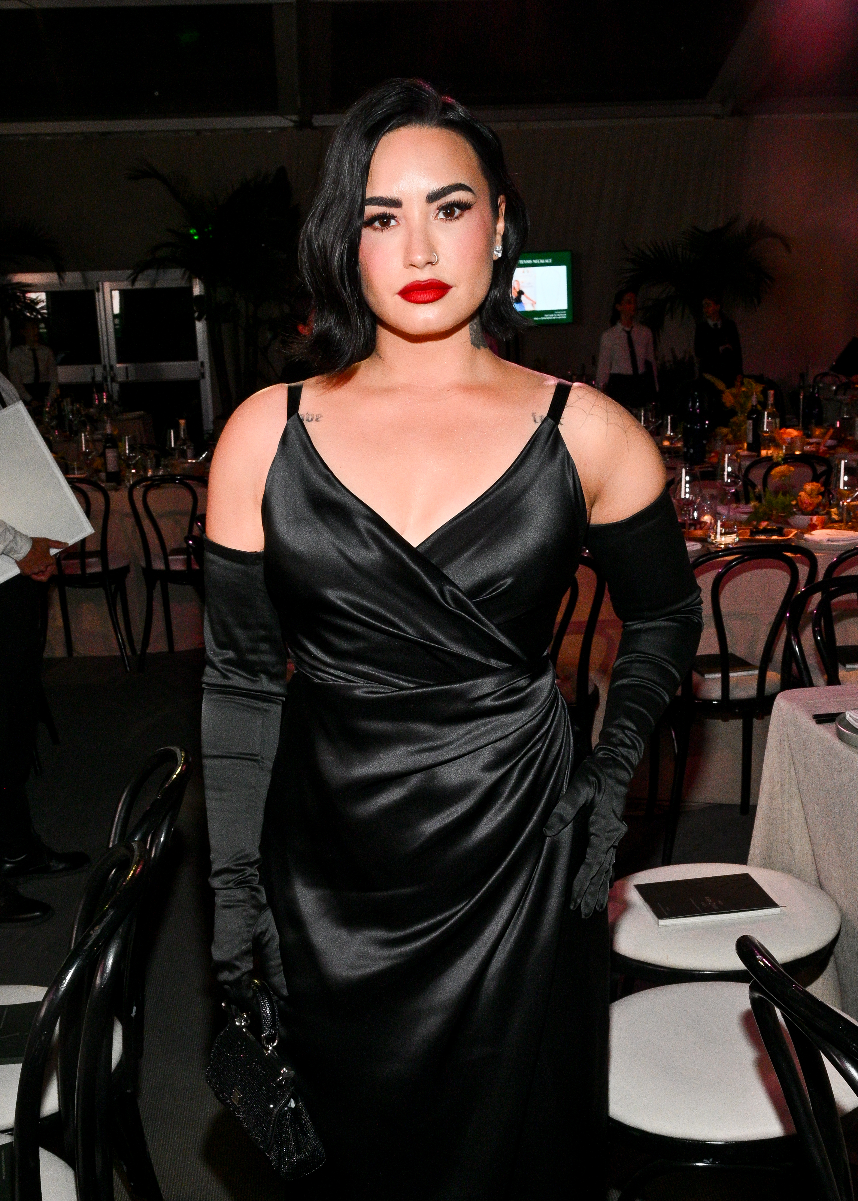 Demi Lovato stands at an event wearing a black, off-the-shoulder satin gown with matching gloves