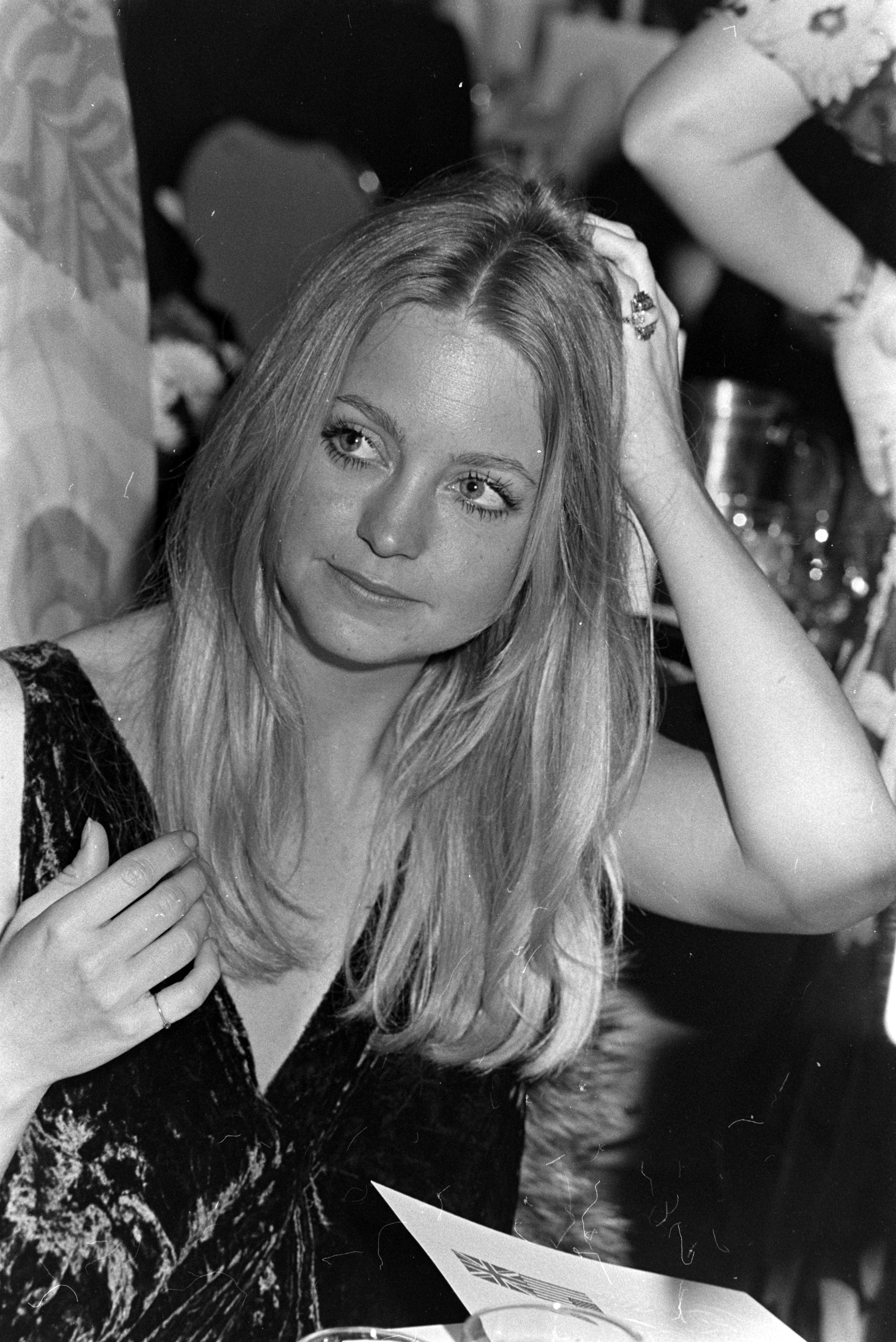 Goldie Hawn seated at an event, adjusting her hair and wearing a sleeveless dress