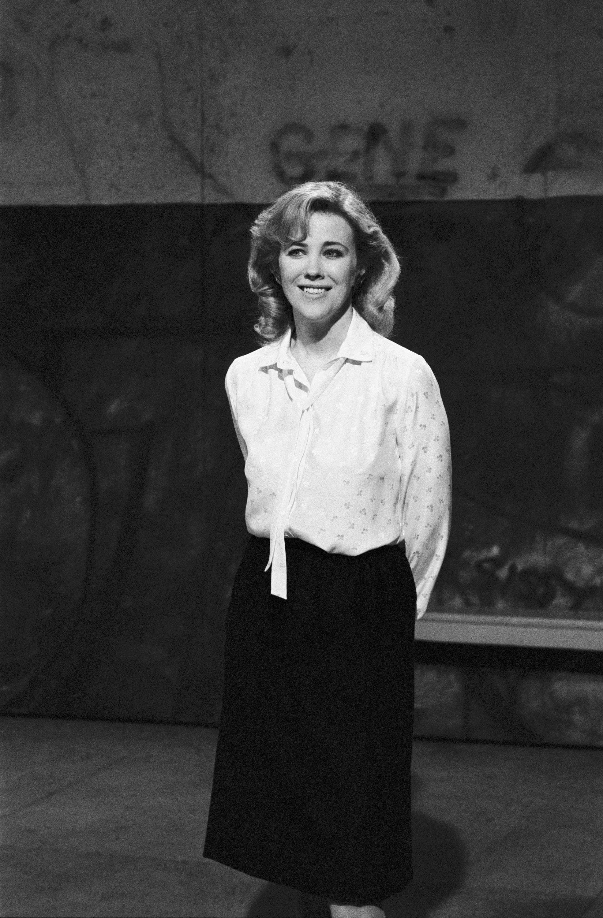 Judy Garland is standing in front of a graffitied wall, wearing a light blouse and dark skirt, smiling