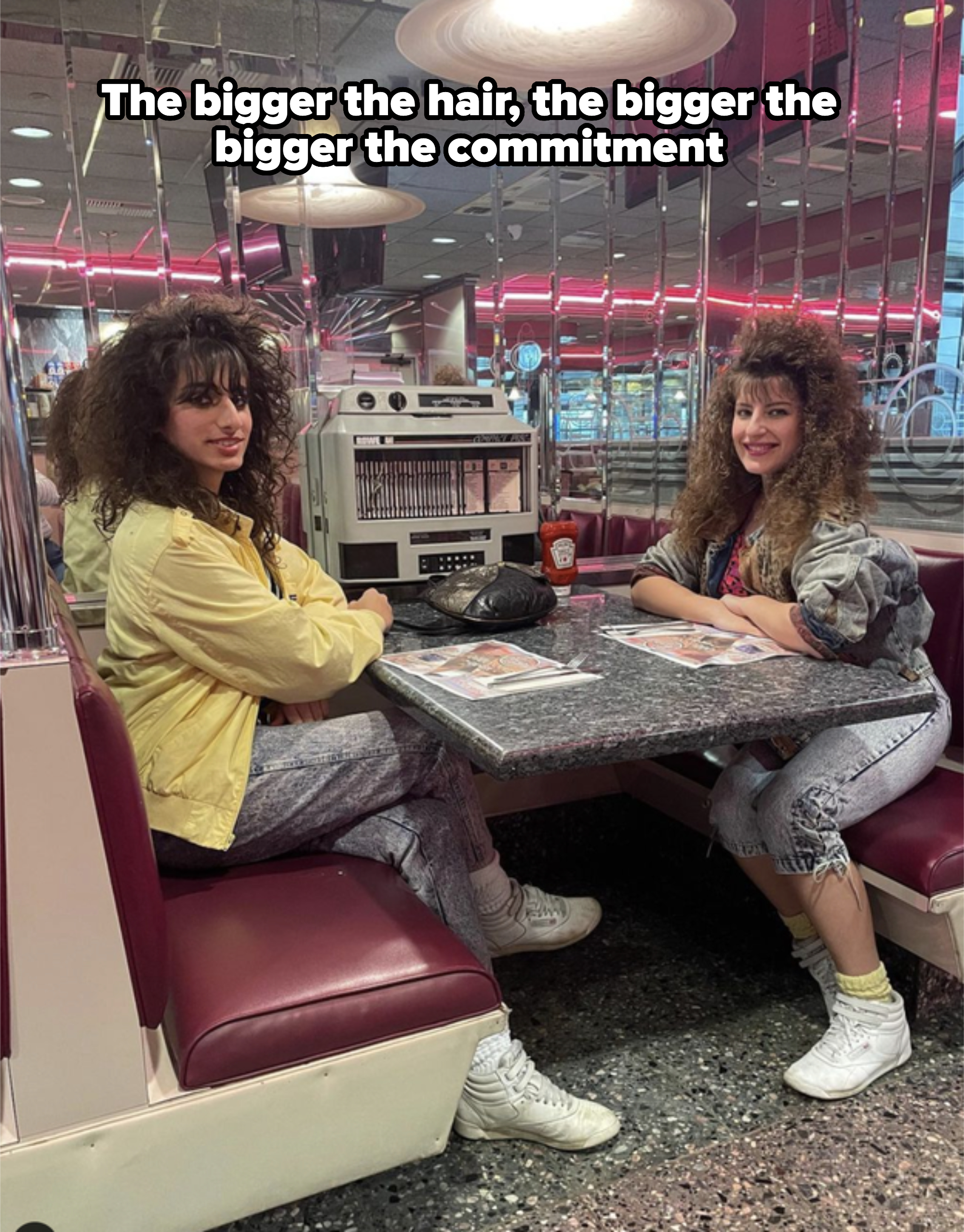 Two people sit at a retro diner booth with mirrors in the background, wearing vintage 1980s-style clothing including jackets and sneakers