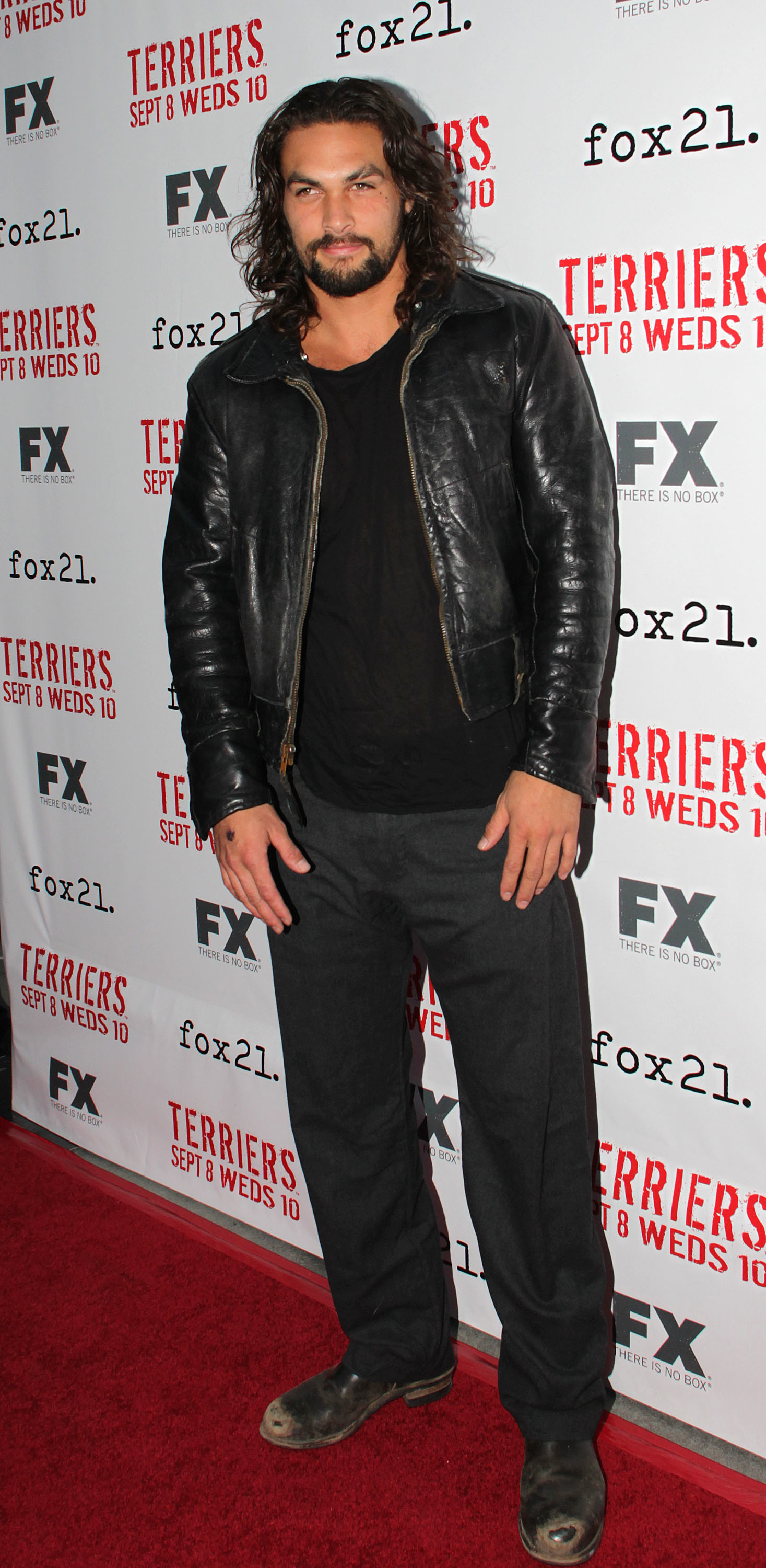Jason Momoa standing on the red carpet, wearing a black leather jacket, black shirt, and dark pants, at an FX "Terriers" premiere event