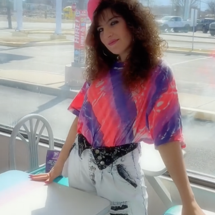 A woman with curly hair wears a vibrant tie-dye shirt, white jeans, and a black belt. She poses in a sunlit diner with large windows