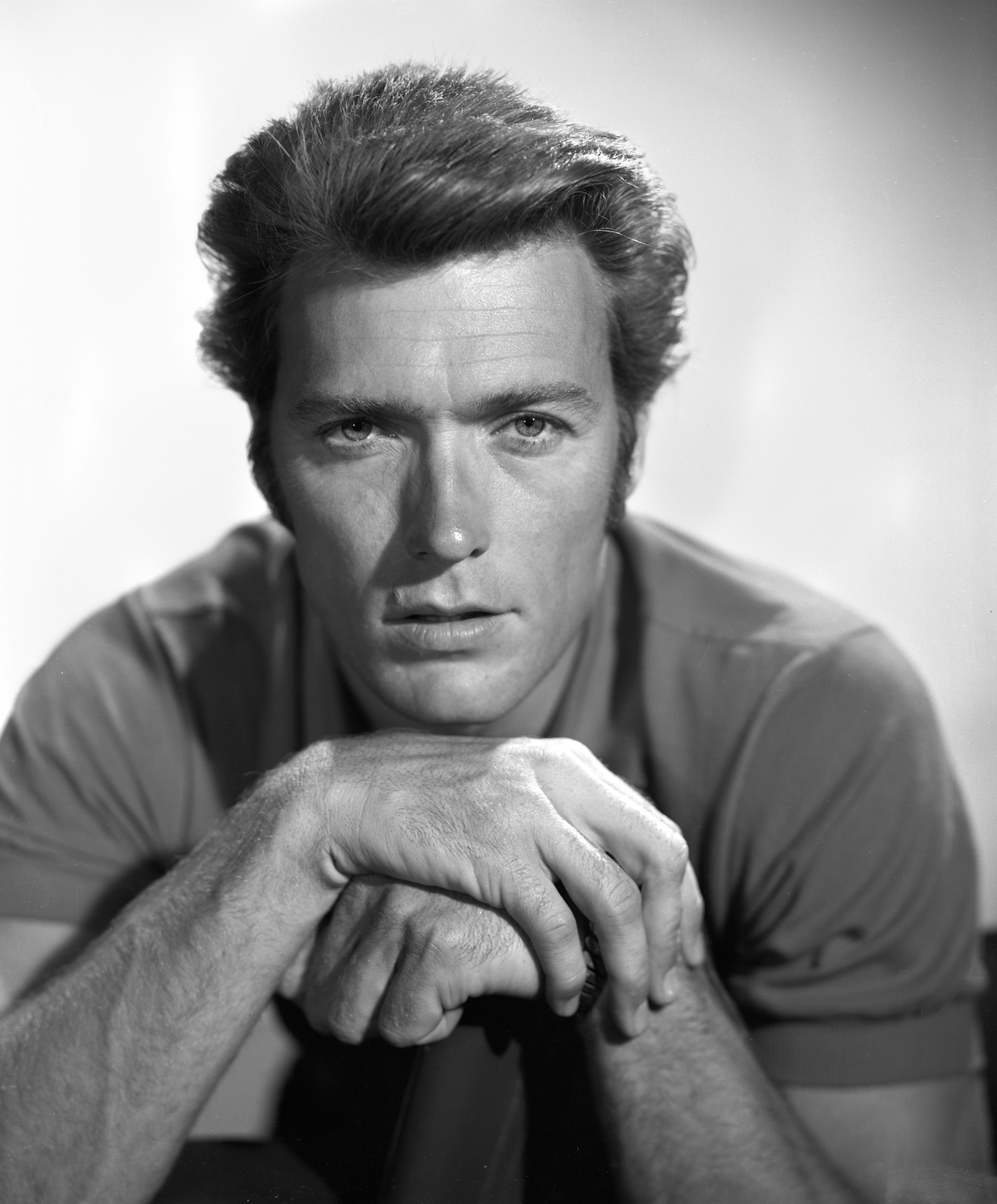 Black and white portrait of Clint Eastwood, looking directly at the camera with hands clasped in front, wearing a simple shirt