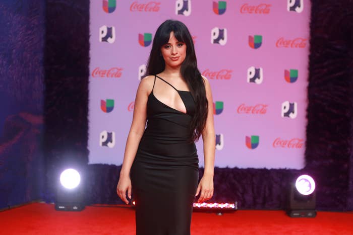 Camila Cabello stands on a red carpet wearing a sleek, black dress with a diagonal strap. The backdrop features logos for Univision and Coca-Cola