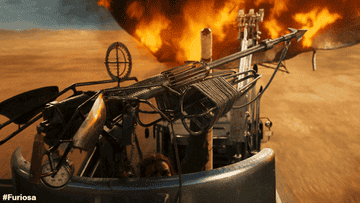A close-up of a high-speed chase scene from "Furiosa" featuring an elaborate vehicle with a mounted weapon, large flames in the background