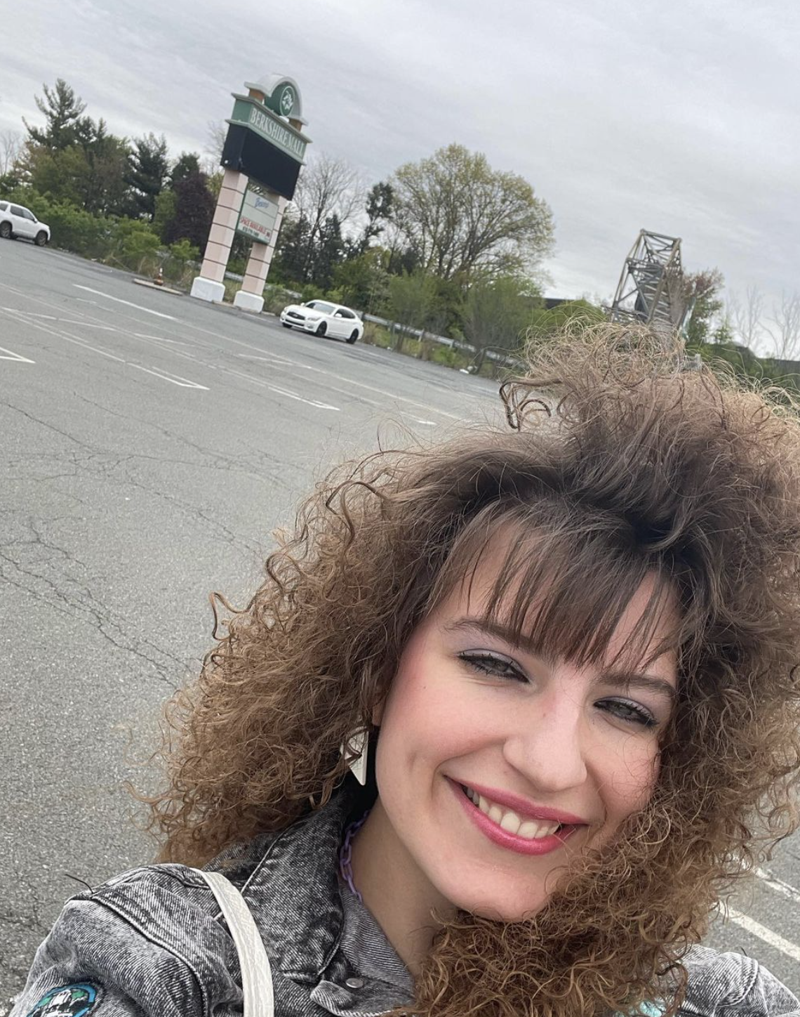 Woman smiling with curly hair and bangs in a nearly empty parking lot with trees and a white car in the background
