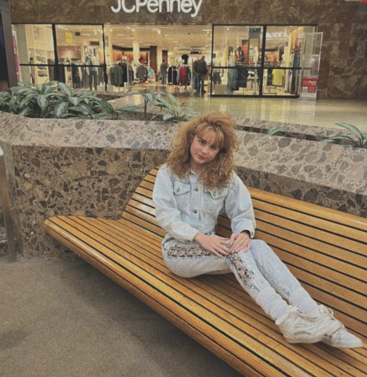 A person with curly hair sits on a wooden bench in front of a JCPenney store inside a mall. They wear a jean jacket, light-colored pants, and sneakers