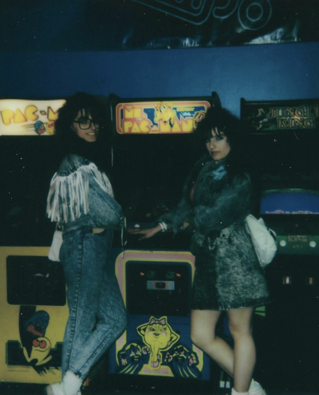 Denise Mercedes and Maria Castellanos pose in front of retro arcade game machines including Ms. Pac-Man and Pac-Man, wearing patterned denim jackets