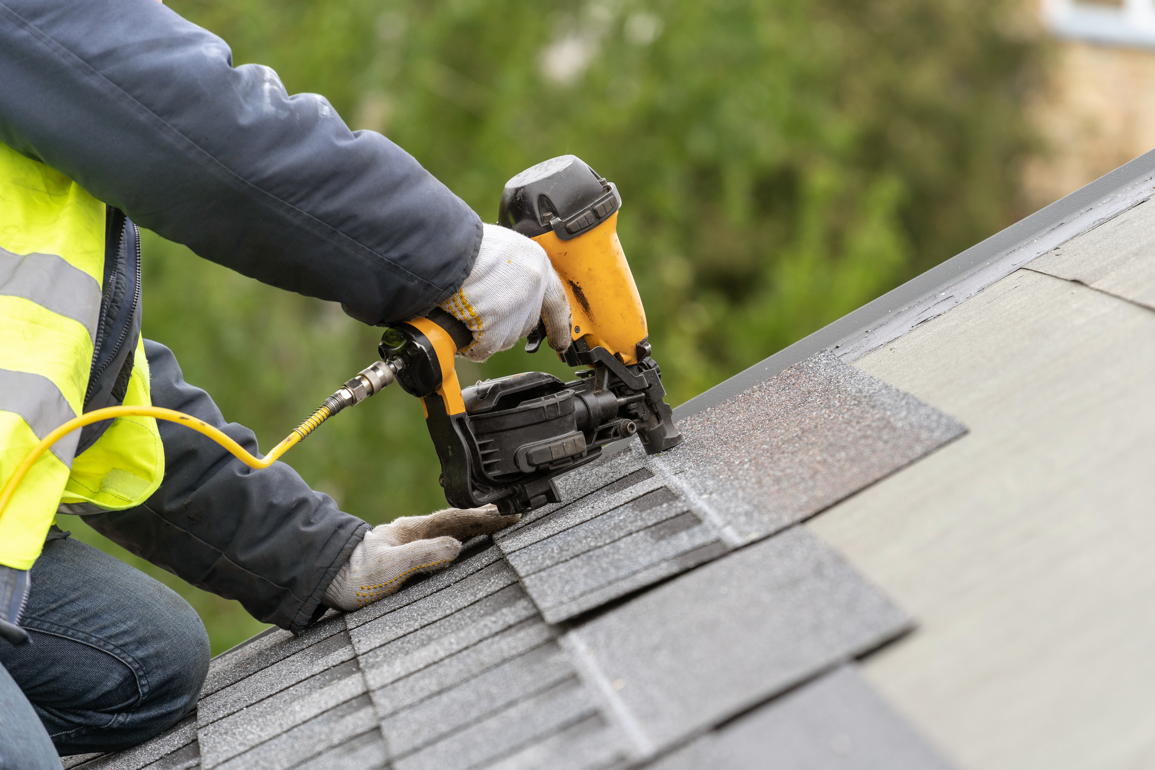 Person using a nail gun to install roof shingles on a house. The person is wearing gloves and a safety vest