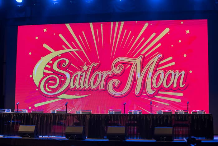 Stage setup with tables, microphones, and chairs in front of a large screen displaying &quot;Sailor Moon&quot; with starburst design. No people are present