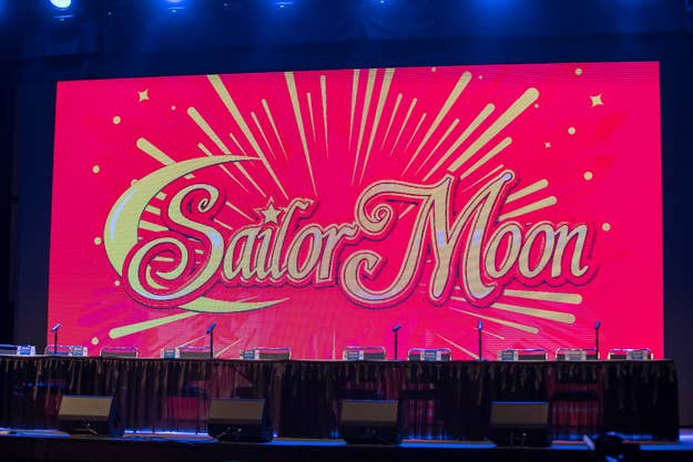 Stage setup with tables, microphones, and chairs in front of a large screen displaying "Sailor Moon" with starburst design. No people are present