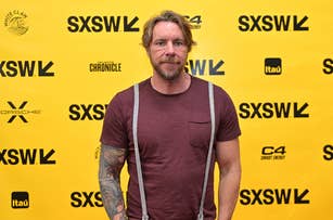 Man with tattooed arms and beard wearing a t-shirt and suspenders on a SXSW event backdrop