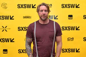 Man with tattooed arms and beard wearing a t-shirt and suspenders on a SXSW event backdrop