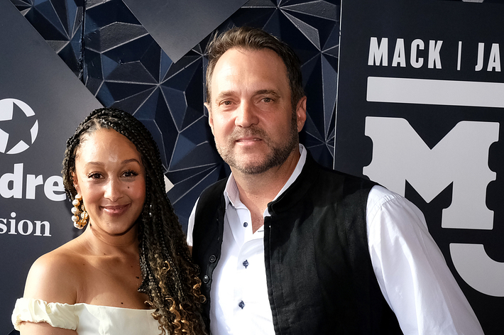 Tamera Mowry-Housley, in an off-shoulder buttoned dress, and Adam Housley, in a vest over a white shirt, pose together on a red carpet