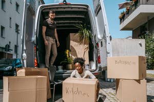 A man and a woman are unloading labeled cardboard boxes from the back of a moving van. Boxes are labeled "Kitchen" and "Bathroom."